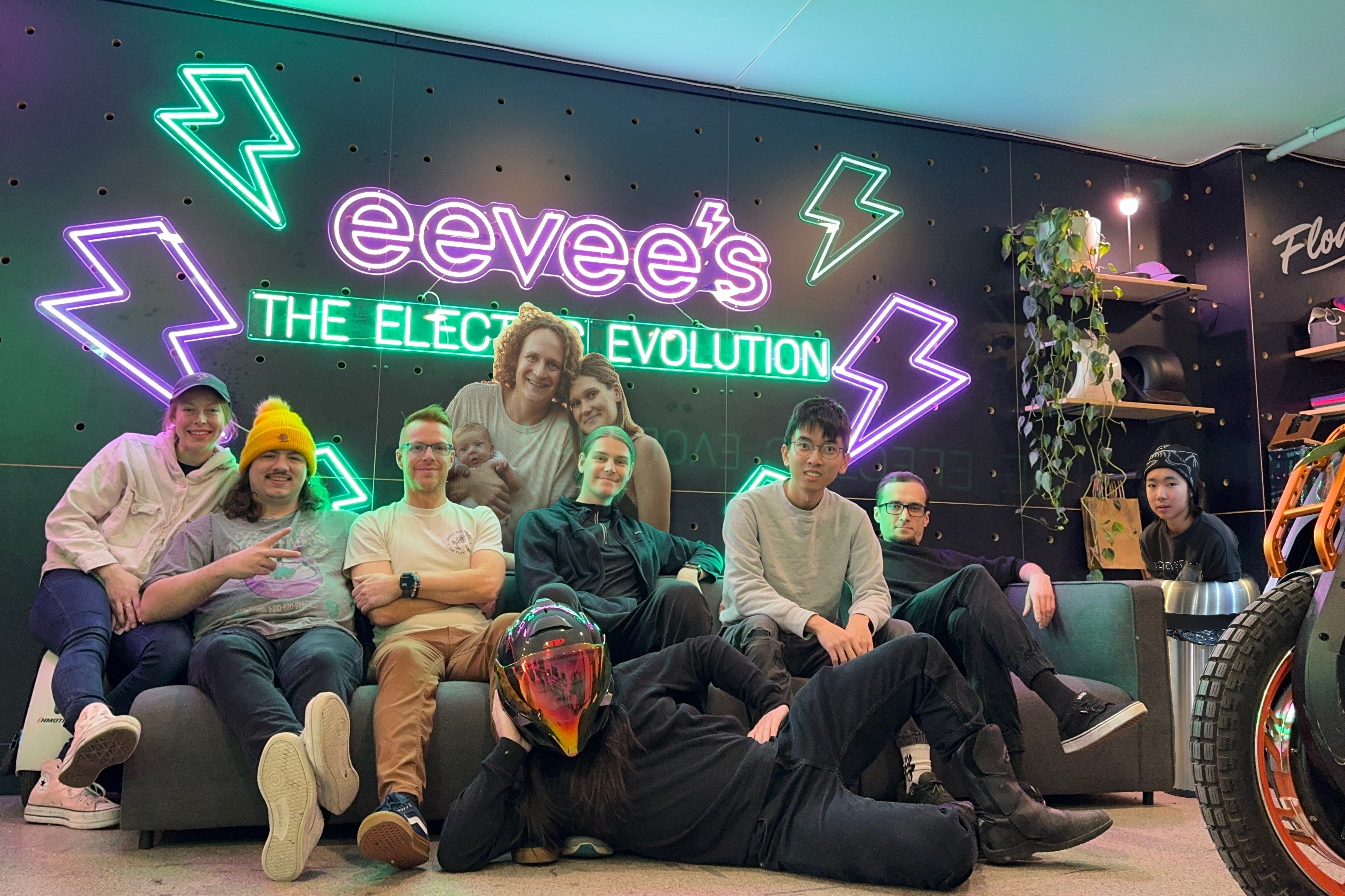 Group of people posing in front of a neon sign with 'eevee's' and 'The Electric Evolution' on a dark wall.