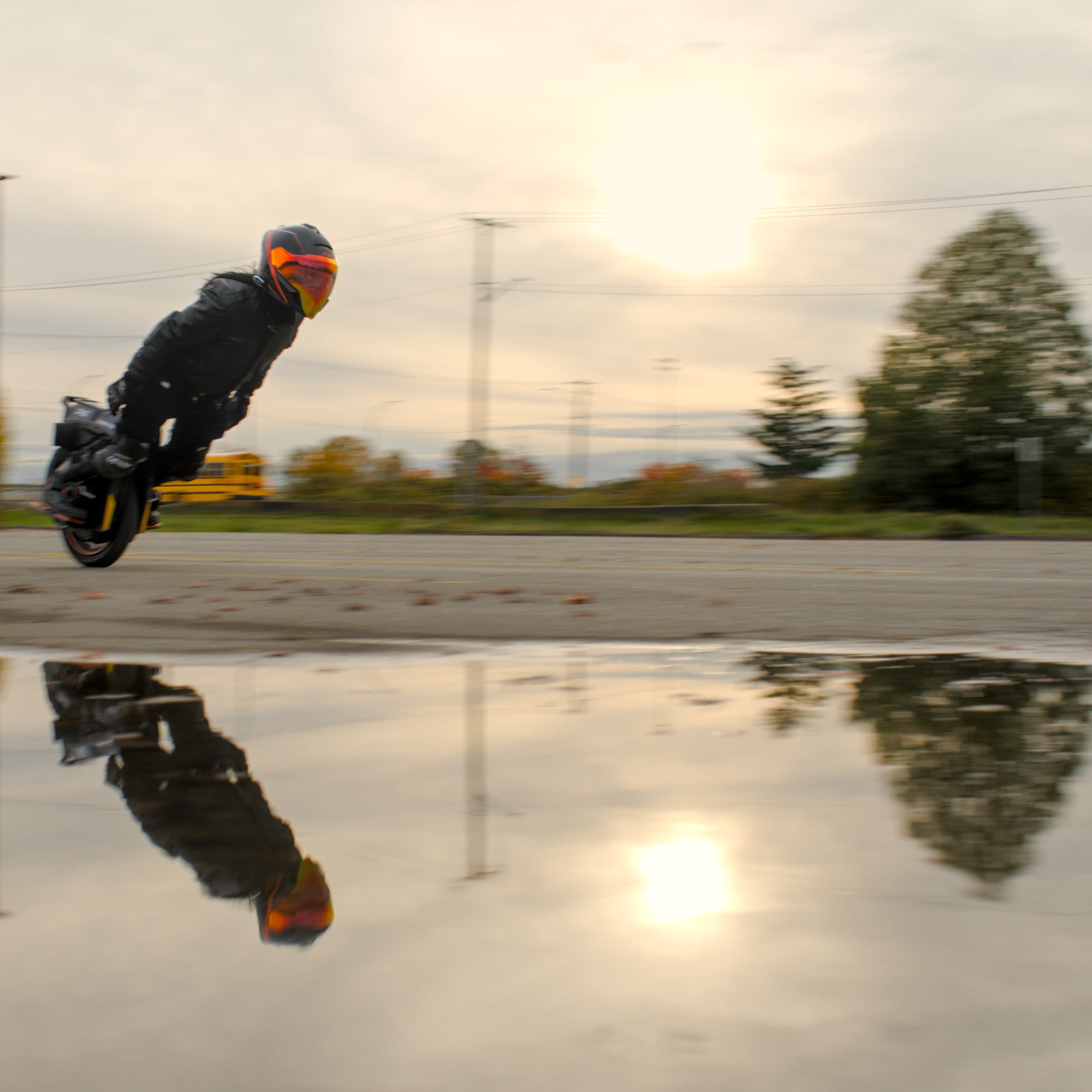 Person performing a heavy lean on an electric unicycle in an open area with trees and power lines in the background.