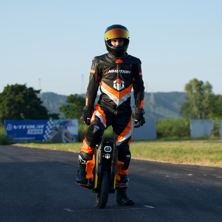 Person in a racing suit and helmet standing on an EUC on racetrack with a clear sky.