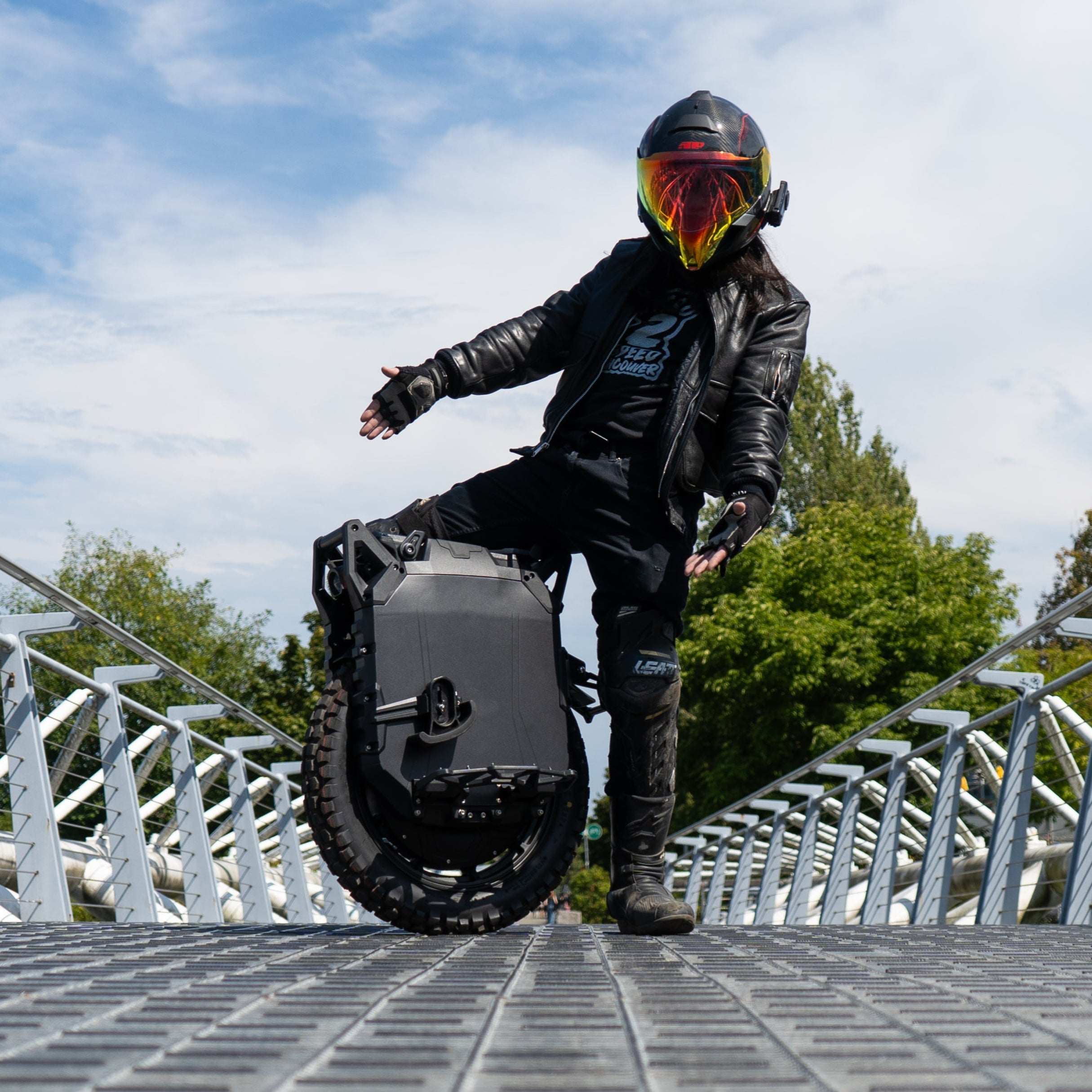 Person riding a unicycle on a metal bridge with a blue sky and trees in the background
